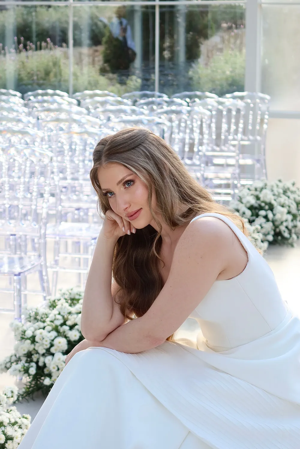 Noiva de vestido branco sentada, com o rosto apoiado na mão, diante de cadeiras de cerimônia e flores brancas.