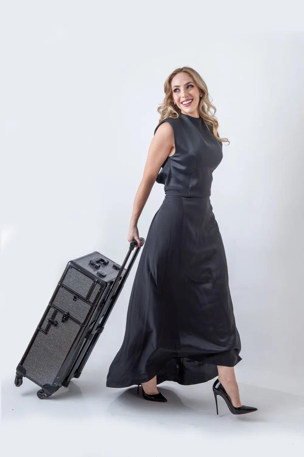 Stella Rotenberg, de vestido preto longo, caminha puxando uma maleta de maquiagem com rodinhas.