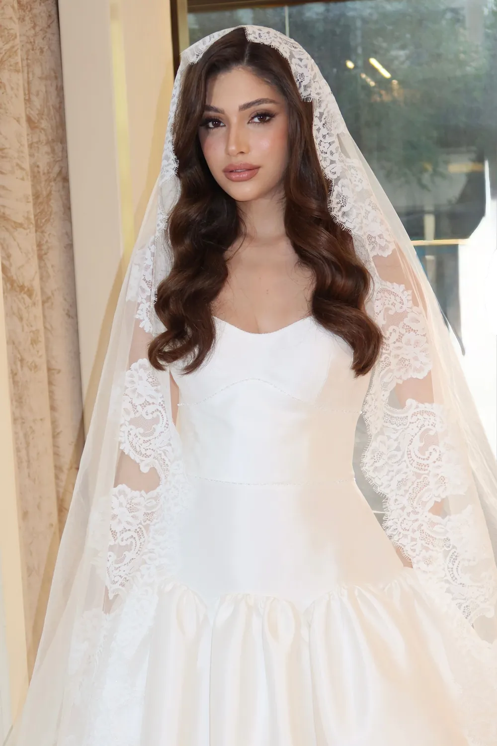 Noiva de vestido tomara que caia e véu longo de renda, em pé diante de uma janela com luz natural.